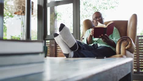 Glücklicher-Afroamerikanischer-älterer-Mann,-Der-Sich-Mit-Erhobenen-Füßen-Im-Sessel-Entspannt-Und-Ein-Buch-Liest