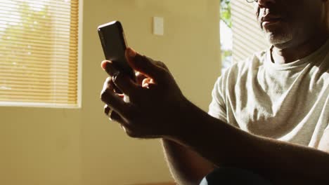 Nachdenklicher-Afroamerikanischer-älterer-Mann,-Der-Zu-Hause-In-Der-Sonne-Sitzt-Und-Sein-Smartphone-Benutzt