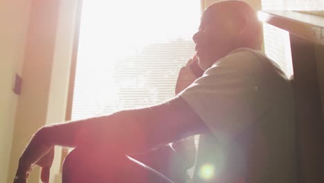 Nachdenklicher-Afroamerikanischer-älterer-Mann,-Der-Zu-Hause-In-Der-Sonne-Sitzt-Und-Auf-Dem-Smartphone-Spricht