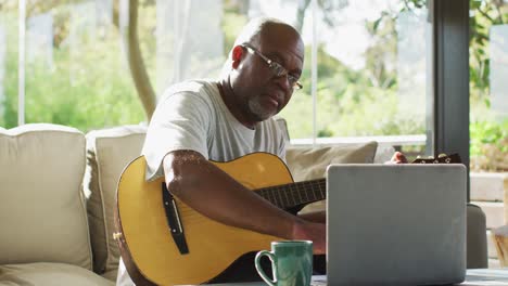 Glücklicher-Afroamerikanischer-älterer-Mann-Im-Wohnzimmer,-Der-Akustikgitarre-Spielt-Und-Einen-Laptop-Benutzt