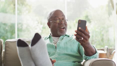Happy-african-american-senior-man-sitting-with-feet-up-making-video-call-with-smartphone-and-waving