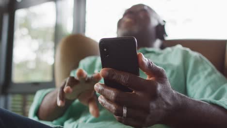 Happy-african-american-senior-man-relaxing-in-armchair,-listening-using-headphones-and-smartphone