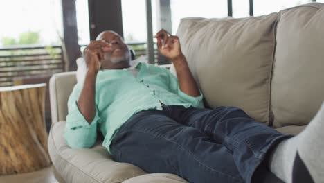 Glücklicher-Afroamerikanischer-älterer-Mann-Entspannt-Sich,-Liegt-Auf-Der-Couch,-Trägt-Kopfhörer-Und-Hört-Musik