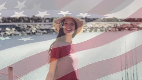 Die-Amerikanische-Flagge-Weht-Vor-Dem-Porträt-Einer-Kaukasischen-Frau-Mit-Hut,-Die-Am-Strand-Lächelt