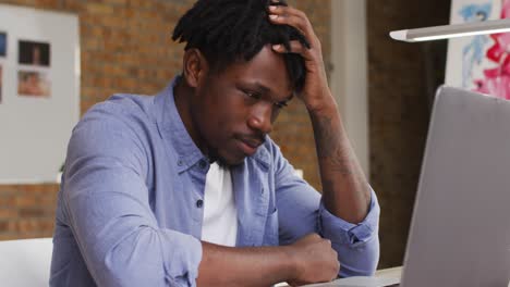 Stressed-african-american-male-artist-looking-at-his-laptop-while-sitting-on-his-desk-at-art-studio