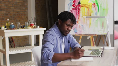 African-american-male-artist-taking-notes-while-sitting-on-his-desk-at-art-studio