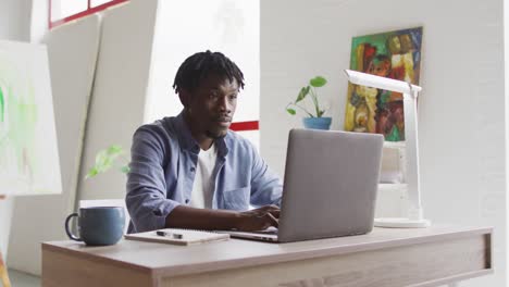 Nachdenklicher-Afroamerikanischer-Männlicher-Künstler,-Der-Einen-Laptop-Benutzt,-Während-Er-Auf-Seinem-Schreibtisch-Im-Kunststudio-Sitzt