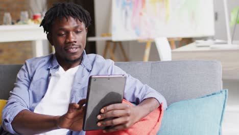 African-american-male-artist-smiling-while-using-digital-tablet-sitting-on-the-couch-at-art-studio