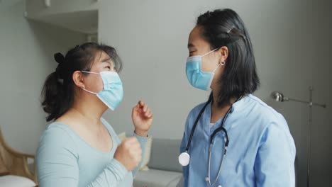 Asian-female-nurse-and-patient-wearing-face-masks-talking-and-looking-to-camera-in-hospital