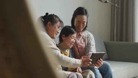 Lächelnde-ältere-Asiatische-Frau,-Die-Ein-Tablet-Benutzt,-Während-Sich-Erwachsene-Tochter-Und-Enkelin-Umarmen