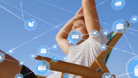 Animation-of-network-of-connections-with-icons-over-man-in-deckchair-on-beach
