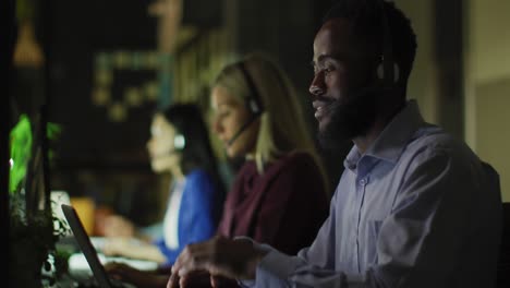 Video-Eines-Lächelnden-Afroamerikanischen-Geschäftsmannes-Und-Seiner-Kollegen,-Die-Nachts-Im-Büro-Headsets-Benutzen