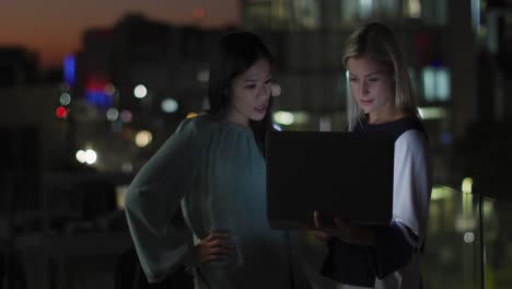 Video-of-two-happy-diverse-businesswomen-talking-and-using-laptop-at-night-in-office
