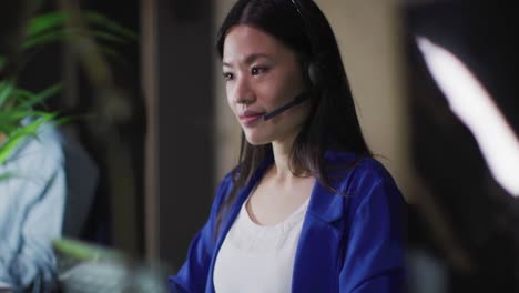 Video-portrait-of-smiling-asian-businesswoman-wearing-phone-headset,-working-at-night-in-office
