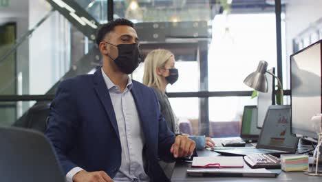 Mixed-race-businessman-wearing-face-mask-and-using-computer-with-colleague-working-in-background