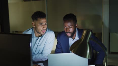Video-of-two-diverse-businessmen-sitting-talking-and-using-a-laptop-together-at-night-in-office