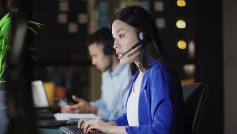 Video-of-asian-businesswoman-talking-on-headset,-working-at-night-with-colleagues-in-office