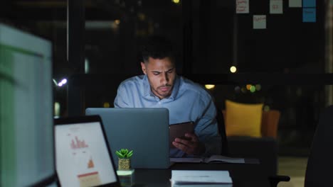 Video-of-biracial-businessman-sitting-at-desk-using-tablet-and-laptop,-working-at-night-in-office