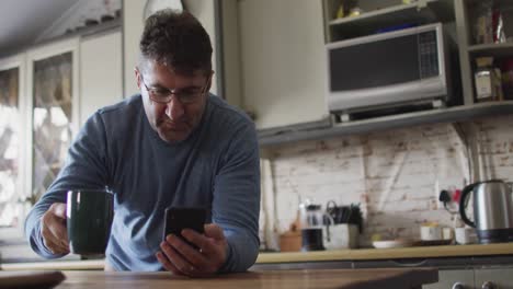 Smiling-caucasian-man-using-smartphone,-drinking-coffee-in-kitchen-at-home