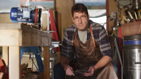 Caucasian-male-knife-maker-sitting-in-workshop,-looking-at-camera-and-smiling