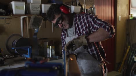Caucasian-male-knife-maker-in-workshop-wearing-glasses-and-headphones-using-sander
