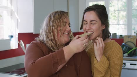Happy-caucasian-lesbian-couple-eating-bread-and-laughing-in-kitchen