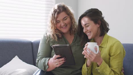 Caucasian-lesbian-couple-using-tablet,-drinking-coffee-and-sitting-on-couch