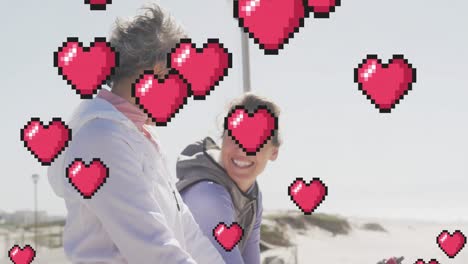 Animation-of-floating-red-pixel-hearts,-over-happy-women-talking-on-beach
