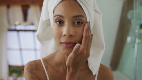 Portrait-of-mixed-race-woman-wearing-towel-on-head-applying-cream-on-her-face