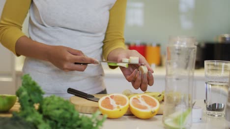 Mid-section-of-mixed-race-woman-preparing-healthy-drink,-cutting-fruit-and-vegetables-in-kitchen