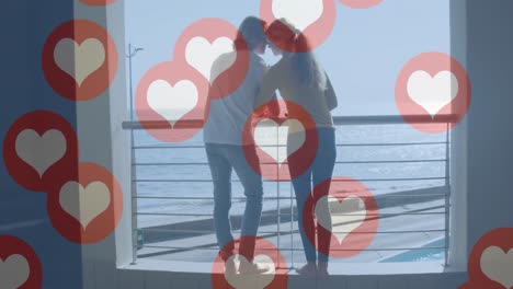 Animation-of-social-media-heart-icons-over-happy-couple-on-balcony-by-seaside