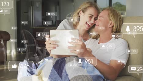 Animation-of-social-media-icons-over-happy-couple-using-tablet-and-embracing-at-home