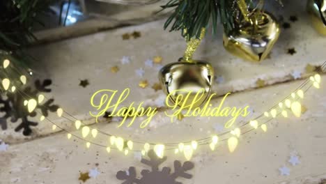 Animation-of-text,-happy-holidays,-in-yellow,-over-string-lights,-bells-and-seasonal-decorations