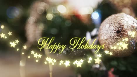Animation-of-text,-happy-holidays,-in-yellow,-over-string-lights-and-blurred-christmas-decorations