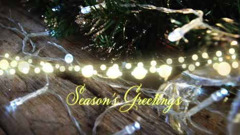 Animation-of-text,-season's-greetings,-in-yellow,-over-string-lights-and-christmas-tree-decorations
