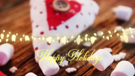 Animation-of-text,-happy-holidays,-in-yellow,-over-string-lights,-marshmallows-and-decorations