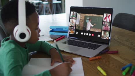 Afroamerikanischer-Junge-Macht-Hausaufgaben,-Während-Er-Zu-Hause-Eine-Videokonferenz-Auf-Dem-Laptop-Hält