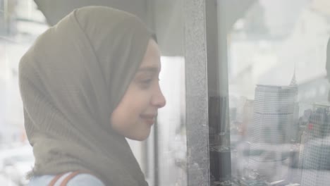 Animation-of-smiling-asian-woman-in-hijab-looking-through-window-over-cityscape