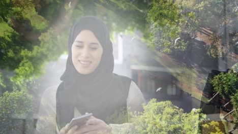 Animation-Einer-Lächelnden-Asiatischen-Frau-Im-Hijab,-Die-Ihr-Smartphone-über-Dem-Wald-Benutzt