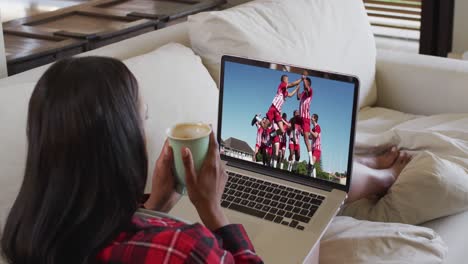 Composite-of-woman-sitting-at-home-holding-coffee-watching-football-match-on-laptop
