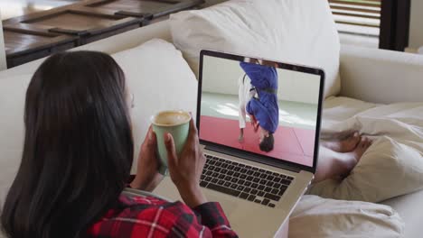 Composite-of-woman-sitting-at-home-holding-coffee-watching-judo-match-on-laptop
