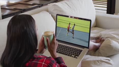 Composite-of-woman-sitting-at-home-holding-coffee-watching-athletics-running-event-on-laptop