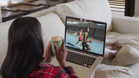 Composite-of-woman-sitting-at-home-holding-coffee-watching-athletics-running-event-on-laptop