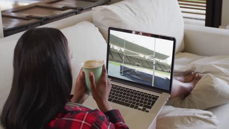 Composite-of-woman-sitting-at-home-holding-coffee-watching-athletics-high-jump-event-on-laptop