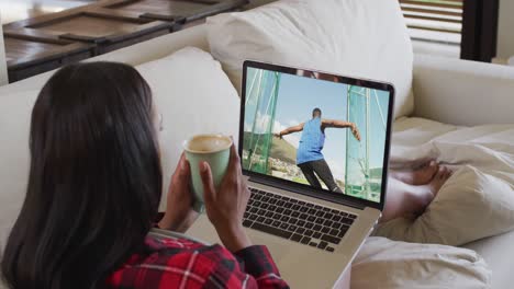 Composite-of-woman-sitting-at-home-holding-coffee-watching-athletics-discus-event-on-laptop