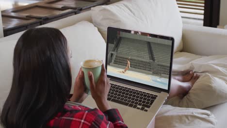 Composite-of-woman-sitting-at-home-holding-coffee-watching-athletics-long-jump-event-on-laptop