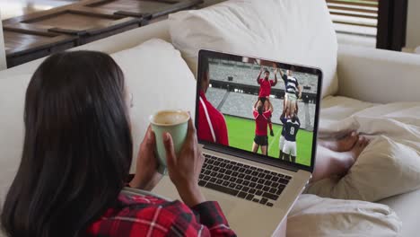 Composite-of-woman-sitting-at-home-holding-coffee-watching-rugby-match-on-laptop