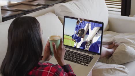 Composite-of-woman-sitting-at-home-holding-coffee-watching-baseball-game-on-laptop