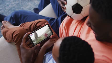 Composite-of-father-and-son-at-home-watching-rugby-match-on-smartphone