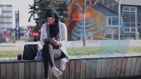 Globe-and-multiple-heart-icons-floating-over-woman-in-hijab-using-digital-tablet-on-the-street